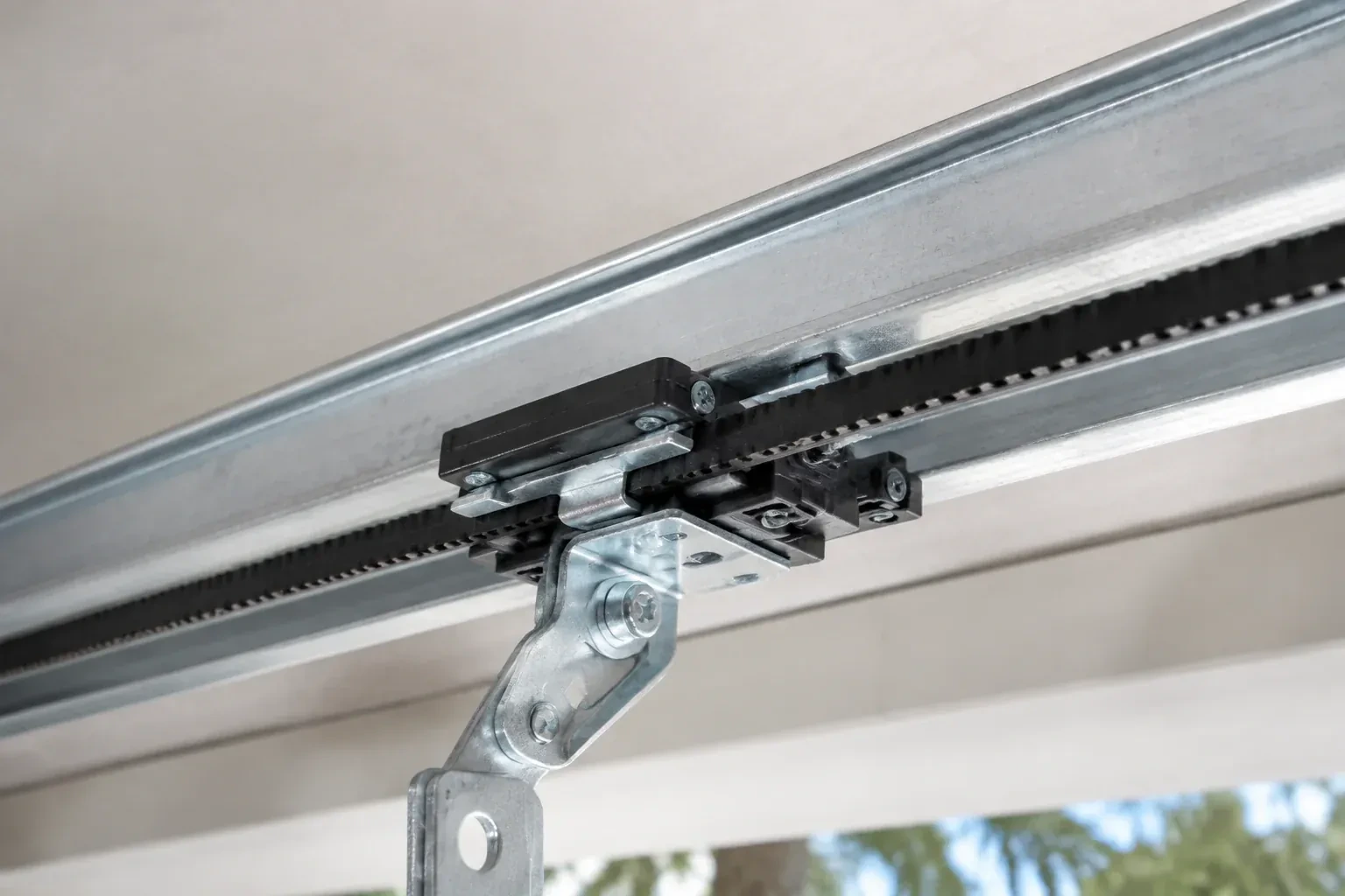 Black plastic trolley and bent galvanized bracket on a steel opener rail with a toothed black belt, against a ceiling. — Belt-Drive Opener Repair, Toronto